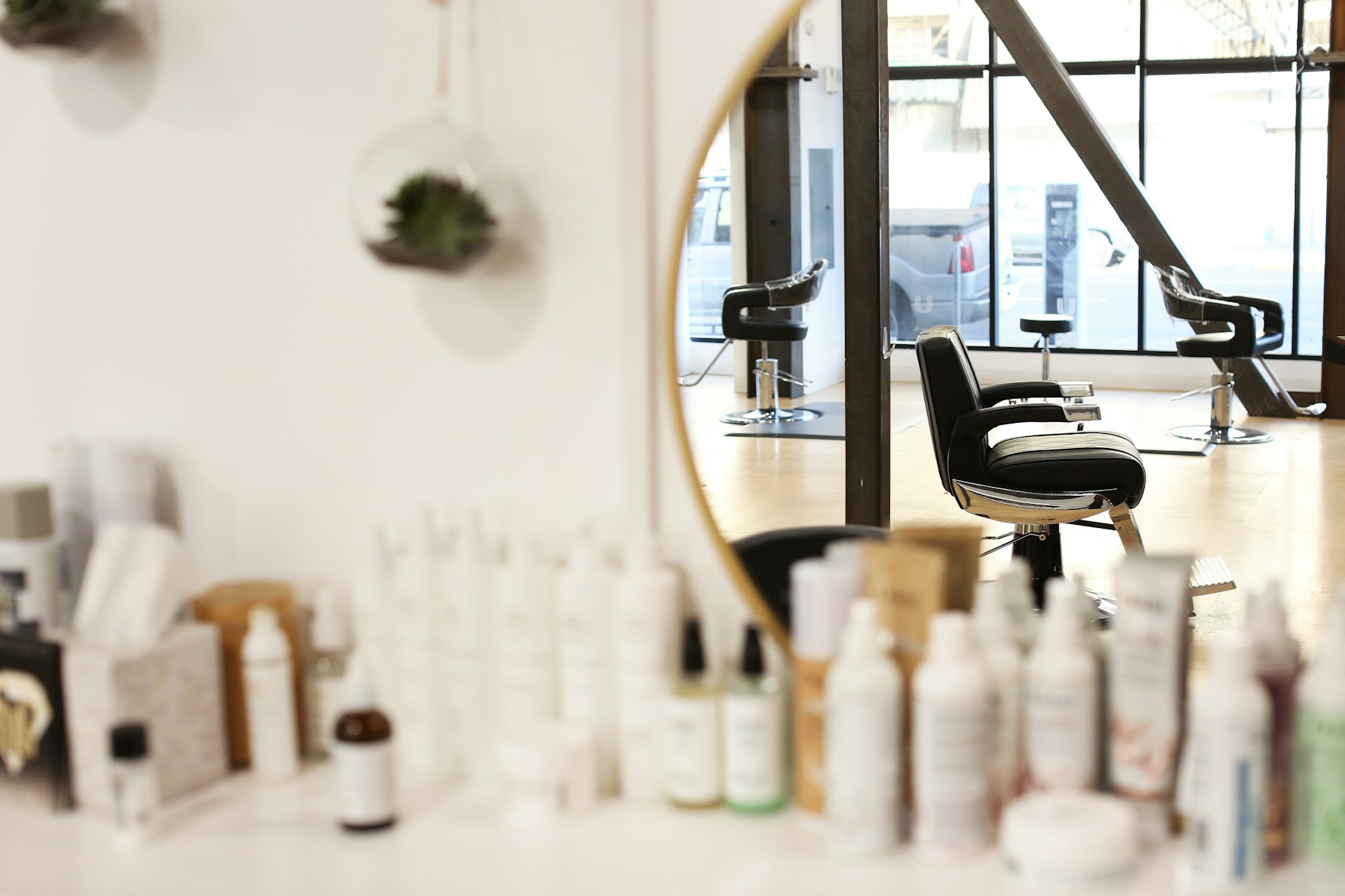 A bright salon with black styling chairs, seen in a round mirror above a shelf of hair products