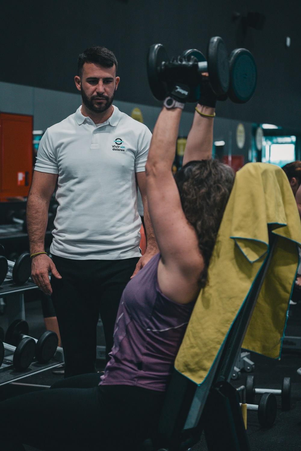 A coach in a white polo watching a client press dumbbells overhead